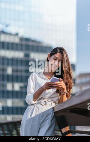 Vertikales Porträt einer attraktiven indischen Frau, die mit ihrem Smartphone eine Sprachnachricht sendet. Sie lehnt sich an das Geländer einer Brücke Stockfoto