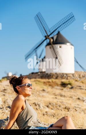 Frau, die auf einem Hügel mit historischen Windmühlen im Hintergrund sitzt Stockfoto