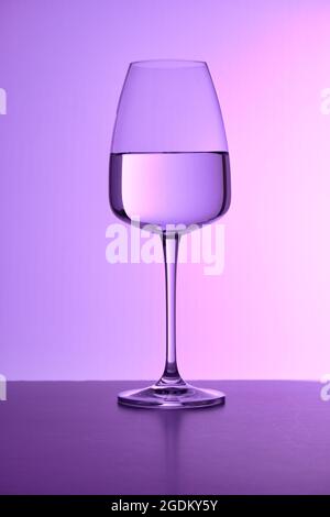 Eine Nahaufnahme eines Glases, das mit reinem Wasser vor dem violetten Hintergrund gefüllt ist. Einfache Oberfläche, keine Spritzer, farbiger Lichteinfall. Restaurant Glaswaren, servieren. Stockfoto