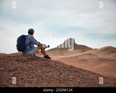 asiatischer männlicher Backpacker Landschaftsfotograf, der auf einem Hügel sitzt und die Aussicht, die Seitenansicht, betrachtet Stockfoto