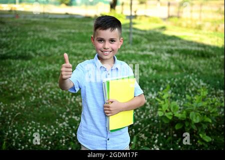 Liebenswert Schuljunge hält ein gelbes Arbeitsbuch und zeigt Daumen bis zur Kamera stehen auf grünem Gras Hintergrund. Glücklicher Elementarschüler kommt zurück t Stockfoto