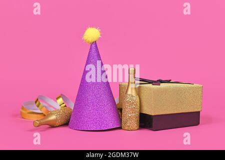 Lila Partyhut, Geschenkbox kleine goldene Champagnerflaschen und Party-Luftschlangen auf rosa Hintergrund Stockfoto