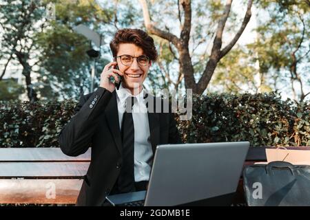 Fröhlicher, professioneller junger Kaukasischer, der auf einer Bank sitzt und seinen Laptop benutzt, um am Telefon zu sprechen Stockfoto