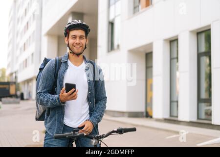 Mittellange Aufnahme eines lächelnden, gutaussehenden männlichen Kuriers mit Thermorucksack, der mit dem Fahrrad auf der Stadtstraße steht und die Navigations-App am Telefon verwendet. Stockfoto