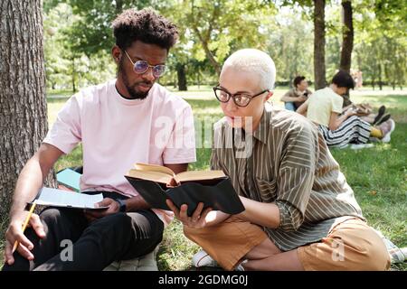 Zwei Studenten sitzen auf dem Rasen und lesen zusammen ein Buch, sie bereiten sich auf Prüfungen im Freien vor Stockfoto