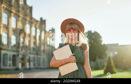 Nettes fröhliches Schulmädchen oder pädagogisches Konzept. Junge blonde Studentin mit Laptop, rosa Sonnenbrille, grünem Kleid und Hut auf dem Universitätscampus. Hochwertige Bilder Stockfoto