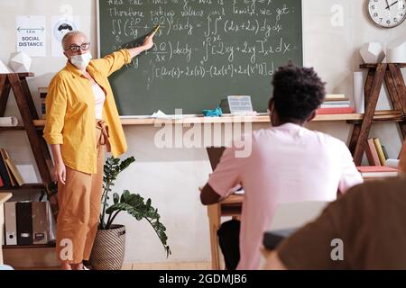 Lehrerin in Maske, die in der Nähe der Tafel steht und auf mathematische Formeln darauf zeigt, erklärt sie den Schülern im Klassenzimmer das Material Stockfoto