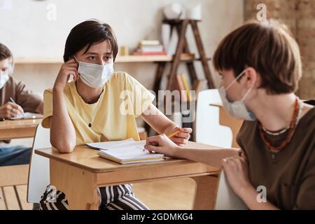 Die Schüler sitzen in Masken an ihrem Schreibtisch und unterhalten sich während des Unterrichts in der Schule Stockfoto