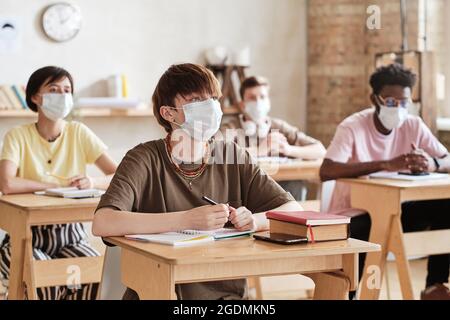 Gruppe von Studenten in Schutzmasken sitzen an Schreibtischen und studieren in der Schule Stockfoto