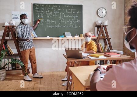 Afrikanischer Student in Maske, der in der Nähe der Tafel stand und darauf zeigte, beantwortete während des Unterrichts die Frage der Lehrer Stockfoto