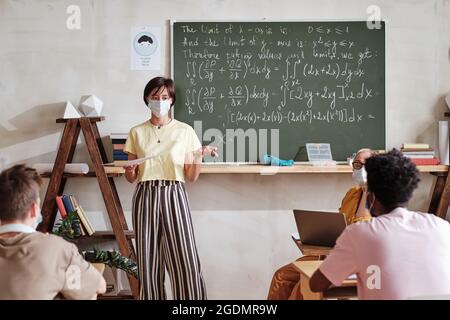 Studentin in Maske, die neben der Tafel steht und eine Prüfung besteht, präsentiert ihren Bericht den Klassenkameraden und dem Lehrer im Klassenzimmer Stockfoto