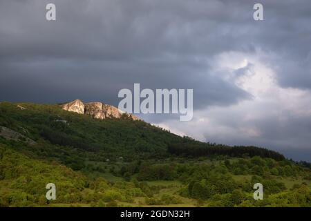 Großartiges, weiches Licht auf einem spitzen, felsigen Berggipfel und einer Landschaft, die von leuchtend grünen, frühlingshaften Wäldern unter einem wolkigen, launischen Himmel bedeckt ist Stockfoto