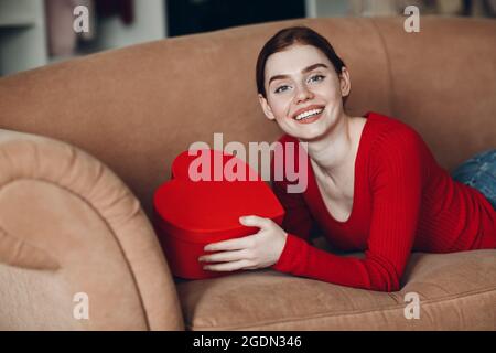 Schöne junge Frau mit roten Haaren, die in ihrem Sofa im Wohnzimmer liegt und die Geschenkbox in Form von Herz und Lächeln hält. Valentinstag Stockfoto