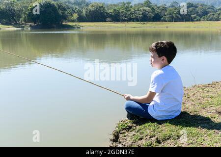 Nahaufnahme eines 8-jährigen brasilianischen Kindes, das an einem sonnigen Morgen am See beim Angeln sitzt. Stockfoto