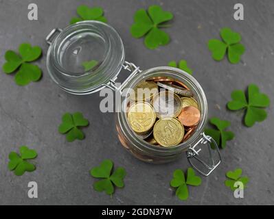 Draufsicht auf Glasglas mit Euro-Münzen auf Schieferplatte mit grünen Kleeblättern Stockfoto