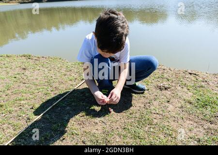 8-jähriges Kind, hockt und legt den Köder auf den Haken, um im See zu fischen. Stockfoto