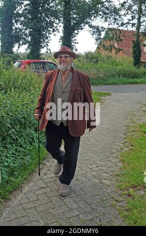 Ein älterer männlicher Wanderer mit einem Stock. Eine braune Jacke und ein brauner Hut in ländlicher Umgebung Stockfoto