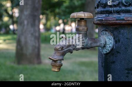 Wasserbrunnen im Park im Sommer, Pumpsystem, Nahaufnahme und Seitenansicht eines schönen Metall-Bronze-Wasserhahn. Teil eines alten Eisenhahns im Freien. Klicken Sie auf Stockfoto