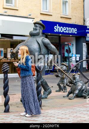 Dundee, Tayside, Schottland, Großbritannien. August 2021. UK Wetter: Ein warmer, luftiger Tag mit viel Sonnenschein in Nordostschottland mit Temperaturen von bis zu 20 Grad Eine junge modische Frau, die in der Nähe der Bronzestatue von DC Thomson der Zeichentrickfigur Desperate Dan steht und Nachrichten auf ihr Mobiltelefon im Stadtzentrum von Dundee schreibt. Kredit: Dundee Photographics/Alamy Live Nachrichten Stockfoto