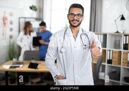 Porträt eines 30-jährigen indischen asiatischen Arztes in einem Klinikraum, der den Daumen hochzeigt und vor der Kamera lächelt, mit zwei fleißigen Kollegen im Hintergrund. Speicherplatz kopieren Stockfoto