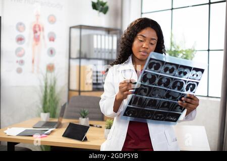 Junge attraktive afroamerikanische Ärztin Radiologin, die das Röntgenbild betrachtet, während sie in einem hellen modernen Büro in der Klinik steht. Allgemeinmediziner, der die Tomographie des Patienten untersucht Stockfoto