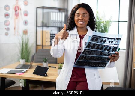 Porträt einer jungen afrikanischen Ärztin mit Stethoskop und MRI-Scan, lächelnd und mit Kamerafrau. Lächelnde Medizinstudentin, die einen Röntgenstrahl hält und den Daumen nach oben zeigt Stockfoto