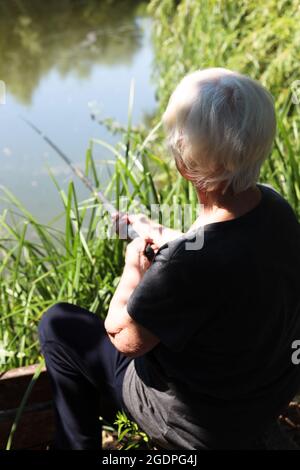 Eine ältere grauhaarige Frau fischt auf dem See. Blick von hinten, hält eine Angelrute in den Händen. Stockfoto