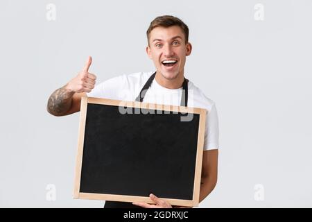 Kleineinzelhandelsinhaber, Café und Restaurant Mitarbeiter Konzept. Enthusiastisch freundlicher und optimistischer Verkäufer in schwarzer Schürze, zeigt Daumen hoch und leer Stockfoto