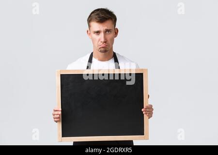 Kleineinzelhandelsinhaber, Café und Restaurant Mitarbeiter Konzept. Enttäuscht und traurig Verkäufer in schwarzer Schürze pouting und unglücklich, zeigen Stockfoto