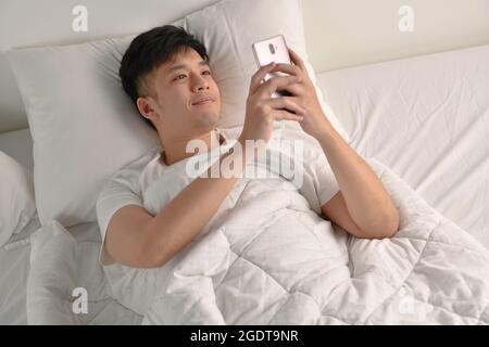 Junger asiatischer Mann, der nach dem Aufwachen am Morgen ein Smartphone im Bett benutzte Stockfoto