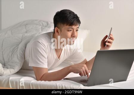 Junger asiatischer Mann, der nach dem Aufwachen am Morgen Laptop und Smartphone im Bett benutzte Stockfoto