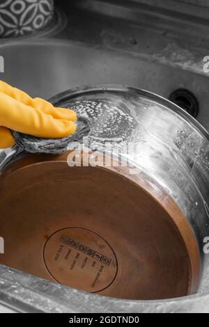 Person, die Gummihandschuhe trägt und einen Kochtopf aus Edelstahl und Kupfer mit einer Drahtunterlage reinigt. Stockfoto