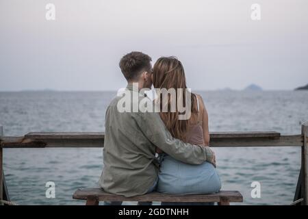 Rückansicht des liebevollen Paares, das auf der Bank in der Nähe des Meeres sitzt. Blick auf den Ozean. Millennials. Romantisches Bild. Hochwertige Fotos Stockfoto