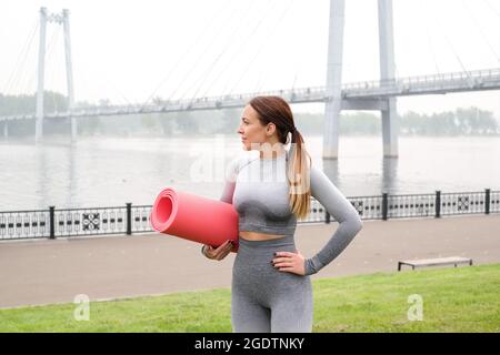 Frau gekleidet Leggings und Top mit rosa Matte tun Yoga im Sommer. Städtischer Hintergrund. Gesundes Sport-Lifestyle-Konzept. Aktive und athletische weibliche verfemte Stockfoto