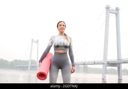 Frau gekleidet Leggings und Top mit rosa Matte tun Yoga im Sommer. Städtischer Hintergrund. Gesundes Sport-Lifestyle-Konzept. Aktive und athletische weibliche verfemte Stockfoto