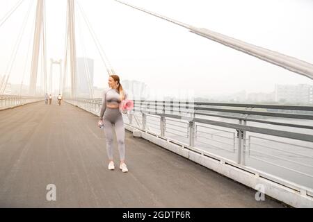 Frau gekleidet Leggings und Top mit rosa Matte tun Yoga im Sommer. Städtischer Hintergrund. Gesundes Sport-Lifestyle-Konzept. Aktive und athletische weibliche verfemte Stockfoto