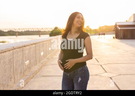 Porträt einer hispanischen Schwangeren, die bei Sonnenuntergang auf dem Damm spazieren geht. Schwangerschaft und Mutterschaft Konzept. Stockfoto