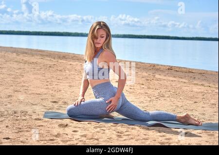 Eine schöne Frau macht Yoga an einem Sandstrand vor dem Hintergrund eines Flusses Stockfoto