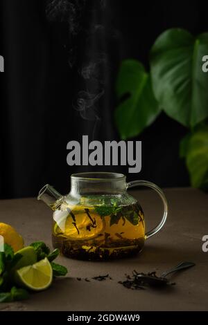 Der Prozess der Zubereitung von schwarzem Tee mit Kräutern, Glas Teekannen mit schwarzem Tee, Zitrone, Limette und Minze in kochendem Wasser getränkt Stockfoto