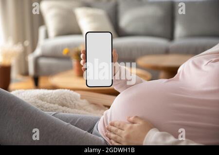 Schwangere zeigt leeren Smartphone-Bildschirm, während sie sich auf der Couch ausruht Stockfoto