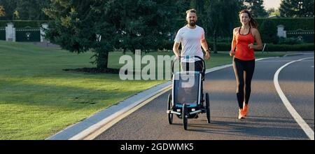 Junge Familie mit ihrem Kind in einem Jogging-Kinderwagen beim Joggen in einem öffentlichen Park. Aktives Familienleben Stockfoto