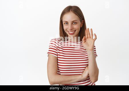 Schöne junge Frau kichert schüchtern, winkt mit der Hand zu Ihnen, sagen Sie Hallo kokett, lächelnd und winken hallo Geste, Gruß jemand, stehend in gestreiftem T-Shirt Stockfoto