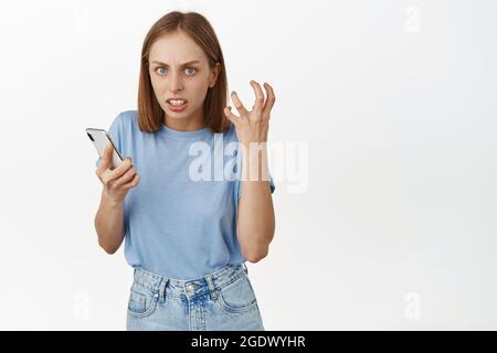 Verärgertes blondes Mädchen, das ihr Mobiltelefon hält, zornig mit der Faust geballt, wütend und irritiert starrt, von jemandem im Internet belästigt wird, hasst soziale Medien Stockfoto