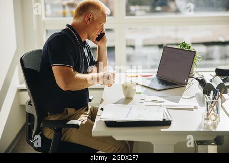 Geschäftsmann, der über das Mobiltelefon spricht, während er vor dem Laptop sitzt. Ein Senior-Mann, der an seinem Schreibtisch sitzt und einen Anruf macht. Stockfoto