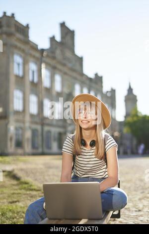 Social Networking, Chatten, Bildung oder Surfen Konzept. Schöne glückliche blonde Schulmädchen, College-oder Universitätsstudentin mit Rucksack und Hut mit Laptop sitzen auf der Bank auf dem Universitätscampus Stockfoto