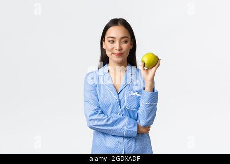 Morgen, aktiv und gesund Lebensstil und Home-Konzept. Schlaue schöne asiatische Frau in blauen Pyjamas Blick nachdenklich auf grünen Apfel, lächelnd schlau Stockfoto