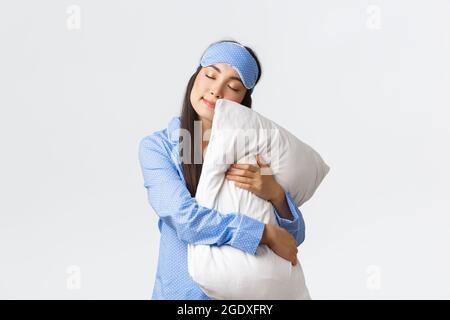 Charmante schlafende asiatische Mädchen in Augenmask und Pyjama, umarmte Kissen und lag auf ihm mit geschlossenen Augen, träumen, mit Nachtruhe, posiert über weiß Stockfoto