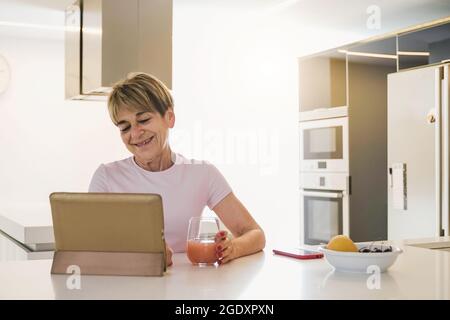 Glückliche ältere Frau, die beim Frühstück in der Küche ein digitales Tablet verwendet – Fokus auf das Gesicht Stockfoto