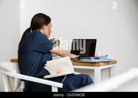 Die Rückansicht einer Ärztin in medizinischer Uniform, die hinter Stroh auf der Suche nach etwas im Laptop im Zimmer sitzt Stockfoto
