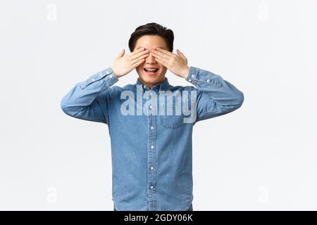 Aufgeregt glücklicher asiatischer Mann mit Zahnspangen, lächelnd amüsiert als Erwartung Überraschung, Hände an den Augen haltend, mit verbundenen Augen stehend, wartet, weiß stehend Stockfoto